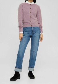 Kvinna klädd i en lavendelknäppt cardigan över en vit skjorta, blå uppvikt jeans och svarta ankelstövlar, stående mot en enkel bakgrund.
