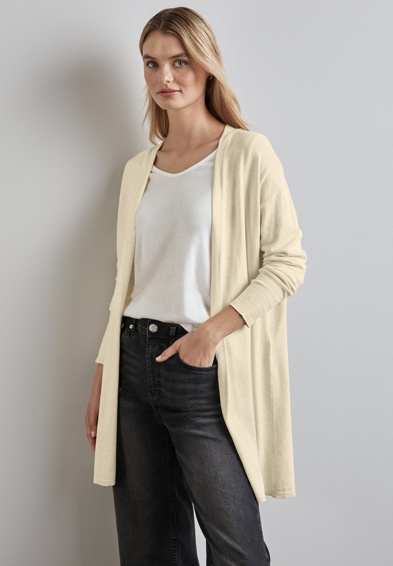 Jasno beżowy długi kardigan o luźnym kroju, noszony na białej koszulce z krótkim rękawem, w połączeniu z czarnymi dżinsami. Miękka tkanina i otwarty przód.