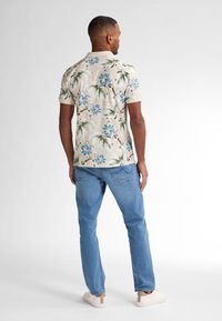 La camicia a maniche corte di colore beige presenta fiori blu e foglie di palma verdi. Indossata con jeans azzurri chiari e scarpe bianche.