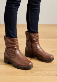 Botas de tobillo de cuero marrón con un eje caído, cremallera lateral, tacón grueso y suela de goma texturizada, combinadas con jeans oscuros sobre un suelo de madera.