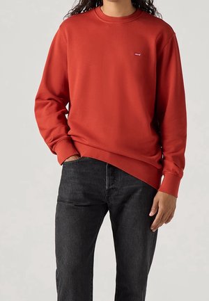 Personne portant un sweat-shirt rouge Levi's et un jean noir, debout avec une main dans la poche devant un fond uni.