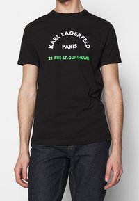 Černé bavlněné tričko s krátkým rukávem. Na přední straně je text "Karl Lagerfeld Paris" v bílé barvě a "21 Rue St-Guillaume" v zelené.
