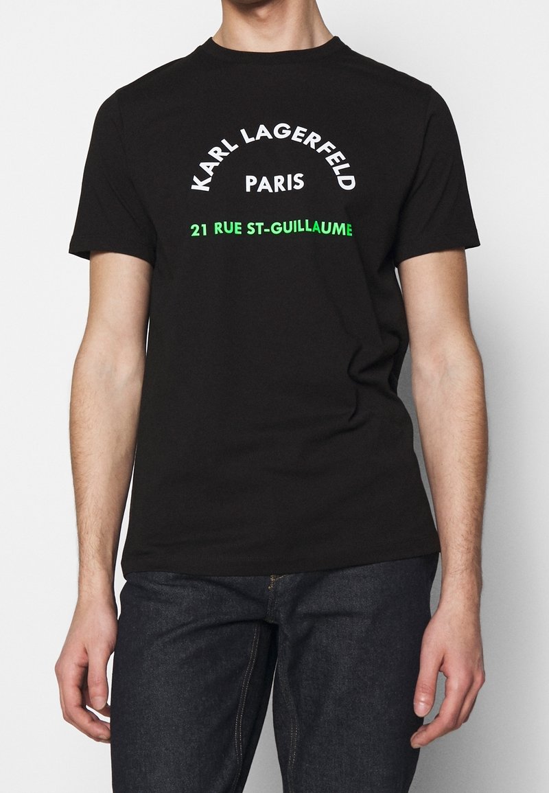 Černé bavlněné tričko s krátkým rukávem. Na přední straně je text "Karl Lagerfeld Paris" v bílé barvě a "21 Rue St-Guillaume" v zelené.