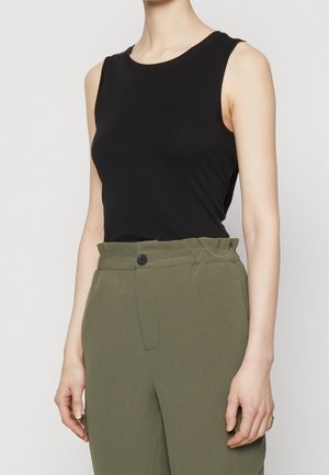 Persona con un top negro sin mangas y pantalones de talle alto en verde oliva con cintura fruncida y cierre con un solo botón.