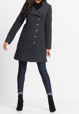 Woman wearing a dark gray buttoned wool coat, dark rolled-up skinny jeans, and black ankle boots standing against white background.