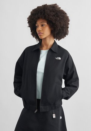 Kvinde med naturligt krøllet hår iført en sort North Face lynlåstrøje over en lyseblå skjorte, stående med hænderne i lommerne, neutral baggrund.