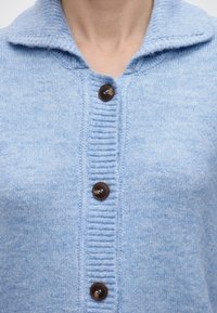 Ανοιχτό μπλε πλεκτό ζακέτα με φαρδύ γιακά και τρία σκούρα καφέ κουμπιά στο μπροστινό μέρος, φορεμένο από ένα άτομο.