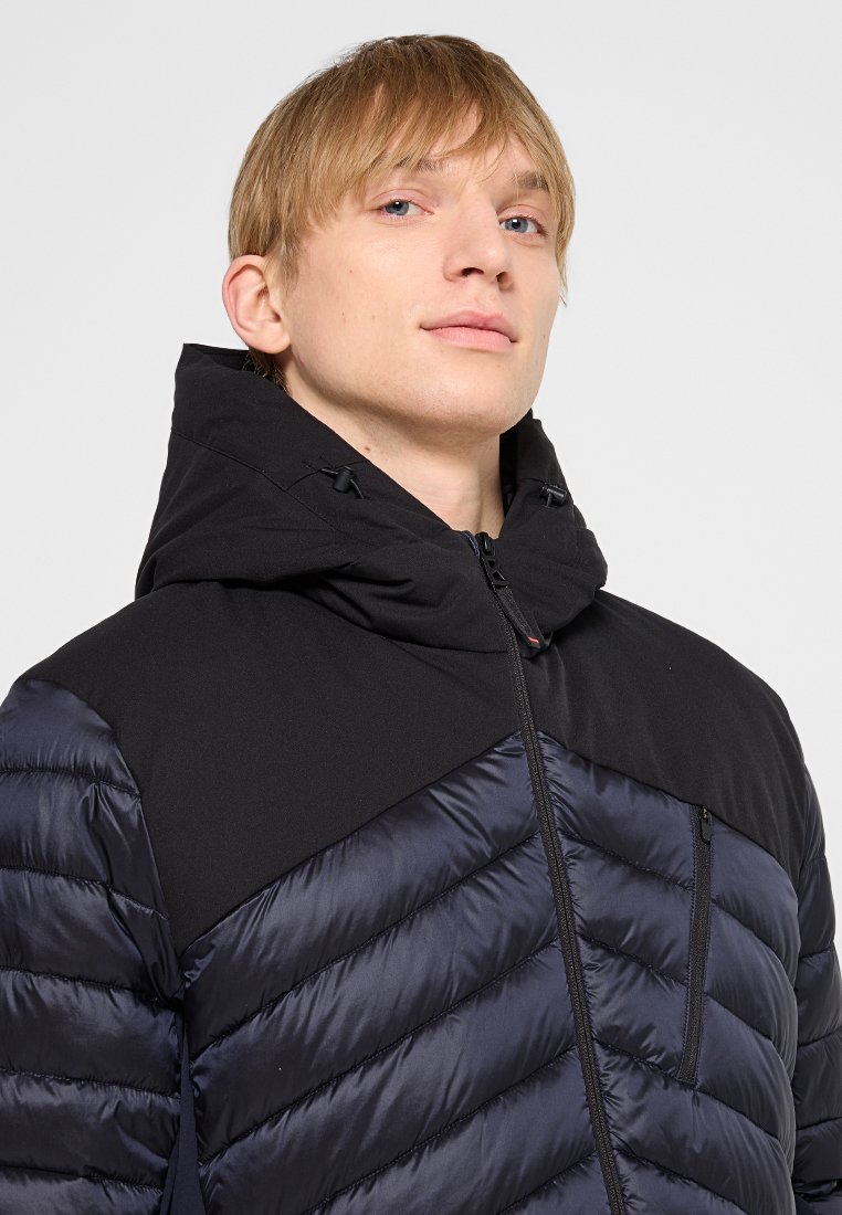 Junger Mann mit kurzen blonden Haaren, der eine schwarze und marineblaue Steppjacke mit hohem Kragen und Kapuze trägt und leicht nach oben blickt.