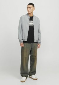 Graue Bomberjacke über einem schwarzen Grafik-T-Shirt, kombiniert mit locker sitzenden dunkelblauen Jeans und weißen Sneakers mit beigen Akzenten.