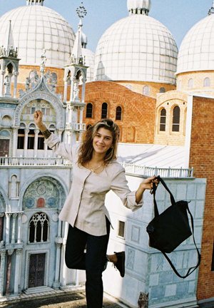 Borsa a spalla nera, camicia button-up beige chiaro, pantaloni scuri, in piedi su un tetto con strutture a cupola e dettagli architettonici ornati sullo sfondo.