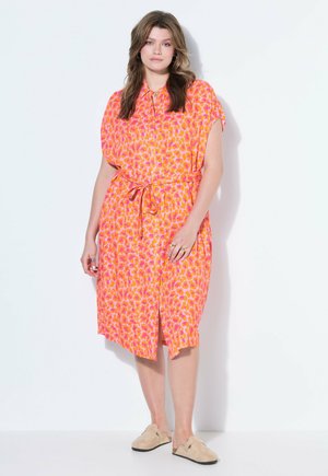 Frau steht an einer weißen Wand, trägt ein knielanges Kleid mit rosa-orangefarbenem Muster, einem Bindegürtel in der Taille und beigen Slip-On-Schuhen.