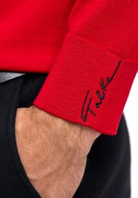 Poignet de pull en tricot rouge avec texture côtelée, arborant le nom de marque brodé en noir, porté avec un pantalon noir.