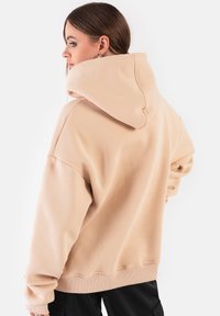 Przesadny beżowy hoodie z teksturowanej tkaniny, z dużym kapturem i opuszczonymi ramionami. Design jest prosty i minimalistyczny.