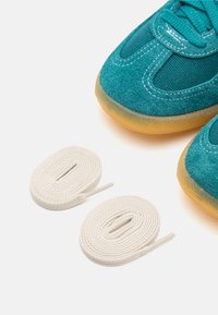 Sapatilhas de camurça verde-água com painéis em malha, solas de borracha e atacadores brancos planos. Close-up dos sapatos e atacadores dispostos sobre um fundo branco.