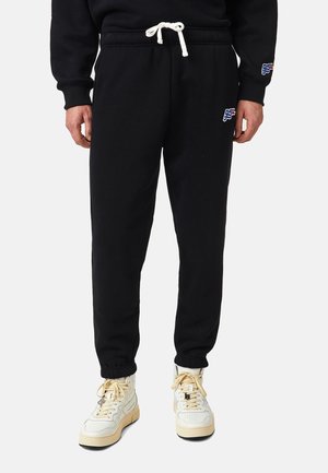 Czarne spodnie dresowe ze sznurkiem w białym kolorze, z małym logo „95L” na lewej udzie i prawym rękawie, noszone z białymi wysokimi trampkami.