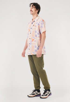 Short-sleeve, button-up shirt with multicolored floral and geometric patterns on a light background; paired with olive green pants and sneakers.