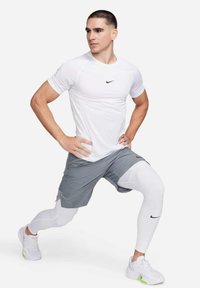 Maglietta atletica bianca a maniche corte con logo nero, pantaloni grigi con design a strati e leggings bianchi. Modello in una posa di affondo.