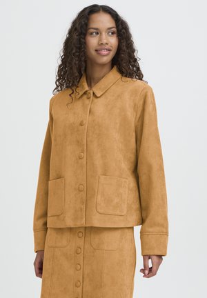 Donna con capelli ricci, indossa una giacca abbottonata color cuoio e una gonna abbinata, guarda leggermente verso sinistra su uno sfondo semplice.