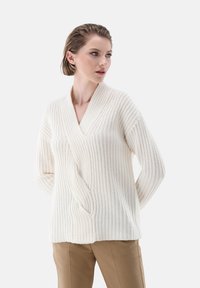 Weißer Strickpullover mit V-Ausschnitt, der ein strukturiertes Muster, lange Ärmel und eine oversized Passform aufweist. Kombiniert mit beigen Hosen.