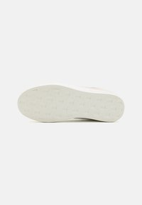 Semelle plate blanche de basket avec un motif subtil en quadrillage et des logos en relief sur un fond clair uni, vue de dessous.