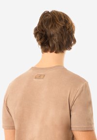Beige T-Shirt mit kurzen �Ärmeln und glatter Textur, ausgestattet mit einem kleinen Markenetikett am Nacken und subtilen Nähdetails.
