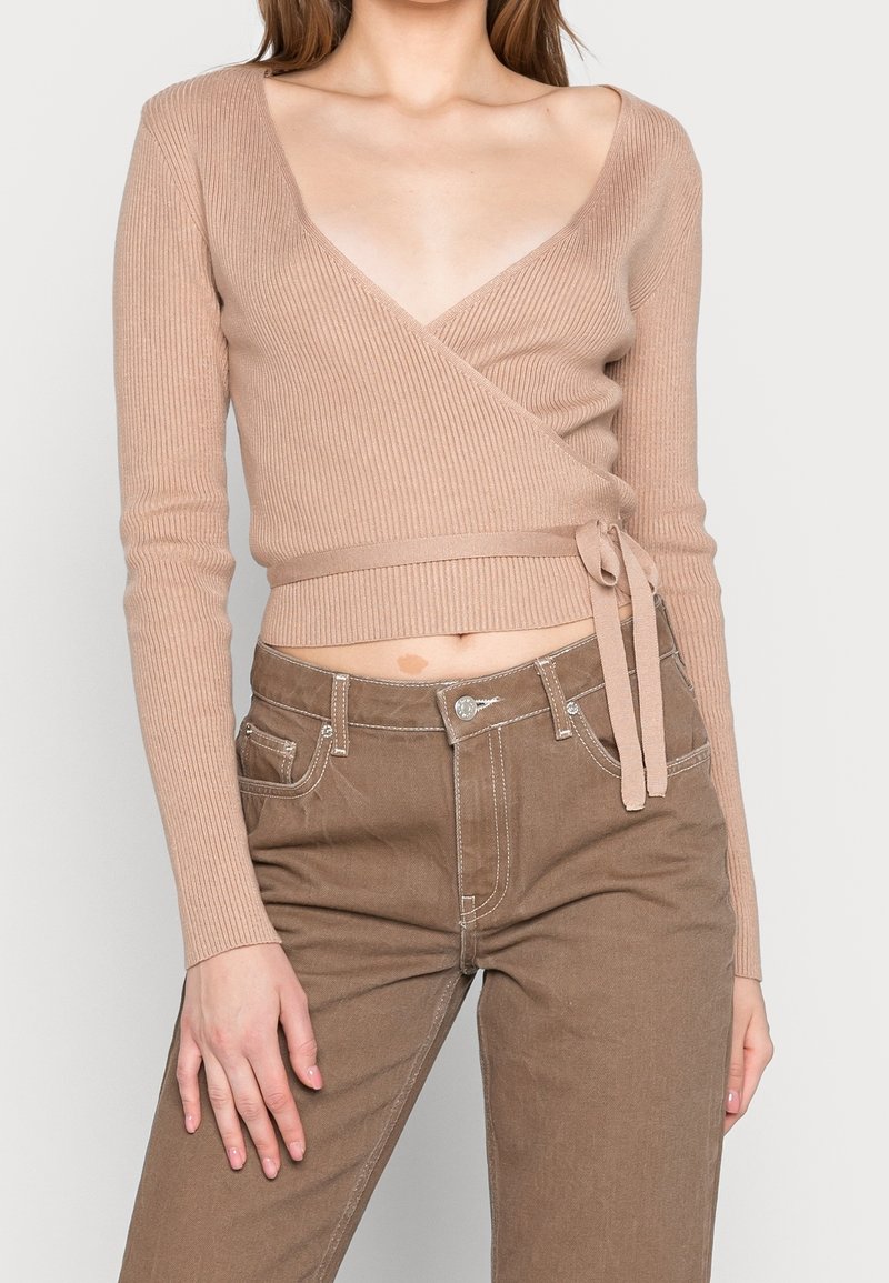 Femme portant un pull cache-cœur côtelé beige noué à la taille avec un pantalon marron taille haute, debout avec une main posée sur la cuisse.