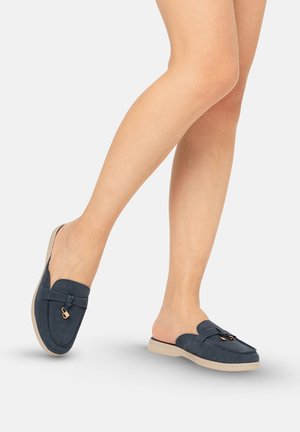 Benen met marineblauwe instap-loafers met beige zolen en kleine gouden gespdetails op een effen lichte achtergrond.