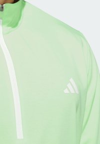 Pullover lungo manica verde chiaro con colletto alto e zip bianca. Presenta un logo bianco sul petto e una texture liscia.