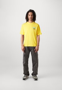 Camiseta de algodón amarilla con mangas cortas y un gráfico en el lado izquierdo del pecho, combinada con pantalones cargo grises que cuentan con múltiples bolsillos y un ajuste relajado.