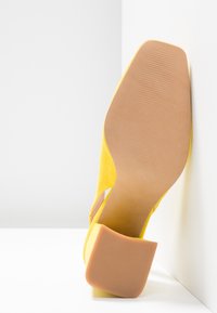 Chaussure à talon en daim jaune reposant sur le côté, montrant une semelle lisse couleur tan et un talon carré sur fond blanc.