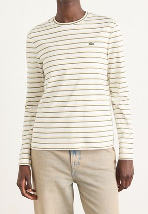 Personne portant une chemise blanche à manches longues avec de fines rayures horizontales jaunes et un pantalon beige sur un fond uni.