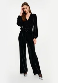 Czarny welurowy kombinezon z długimi bufkami, głębokim dekoltem w kształcie litery V oraz dopasowaną talią związaną pasującym paskiem; szerokie nogawki tworzą zwiewną sylwetkę.