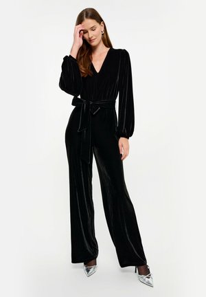 Czarny welurowy kombinezon z długimi bufkami, głębokim dekoltem w kształcie litery V oraz dopasowaną talią związaną pasującym paskiem; szerokie nogawki tworzą zwiewną sylwetkę.