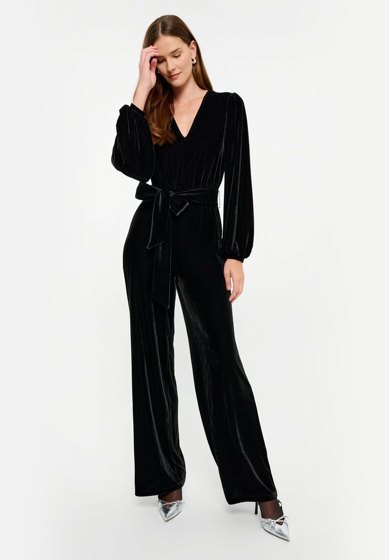 Czarny welurowy kombinezon z długimi bufkami, głębokim dekoltem w kształcie litery V oraz dopasowaną talią związaną pasującym paskiem; szerokie nogawki tworzą zwiewną sylwetkę.