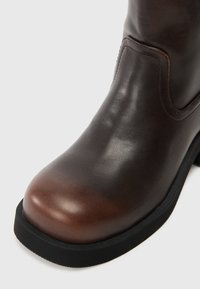 Botă din piele maro cu vârful rotund și talpă din cauciuc negru. Prezintă o textură netedă și cusături de accent în jurul vârfului.