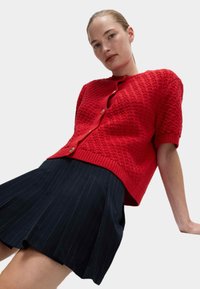 Czerwona dziergana kardigan z teksturowanym wzorem, z krótkimi rękawami i zapięciem na guziki. Połączony z ciemną spódnicą plisowaną w paski.