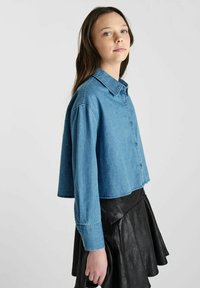 Korte denim shirt in lichtblauw met knoopsluiting, gespreide kraag en textuurdetails, gecombineerd met een zwarte leren rok.