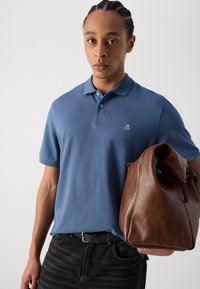 Un tricou polo albastru cu guler și un logo brodat, asortat cu blugi închiși la culoare și ținând o geantă din piele maro. Textură de țesătură netedă.