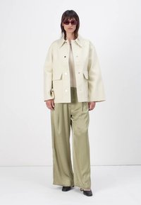 Cream oversized jacket with large pockets, worn over a beige top and olive green wide-leg pants. Accessorized with red sunglasses.