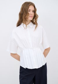 Camisa blanca de manga corta con un diseño de frente plisado, cuello con solapa y tejido suave, combinada con pantalones oscuros.