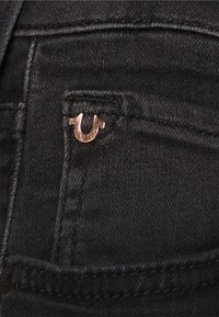 Los jeans de mezclilla negros presentan un pequeño acento de logo metálico en el bolsillo para monedas. La tela parece suave con detalles de costura visibles.