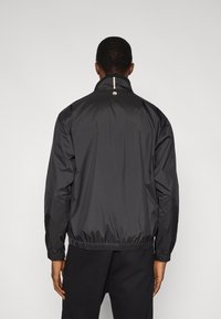 Μαύρο ελαφρύ μπουφάν με ψηλό λαιμό, ελαστικά μανίκια και συγκεντρωμένη λαιμόκοψη. Διαθέτει μικρό λογότυπο στο πίσω μέρος κάτω από το γιακά.