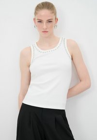 Top bianco senza maniche in tessuto testurizzato, caratterizzato da dettagli in metallo argento lungo il collo. Abbinato a pantaloni neri.