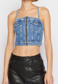 Top corto de mezclilla con cremallera frontal, que cuenta con dos bolsillos en el pecho y tirantes finos. Color azul claro con zonas desgastadas y un ajuste entallado.