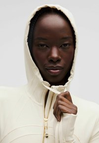 Sudadera cremoso con cremallera hecha de un tejido suave, con detalles en cremallera dorada, que presenta un cuello alto y puños con orificio para el pulgar.