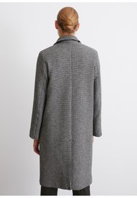 Femme aux cheveux blonds en chignon portant un long manteau à motif pied-de-poule noir et blanc, debout de dos sur un fond blanc uni.
