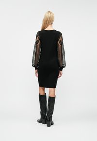 Juoda megzta suknelė su skaidriomis juostuotomis juodomis rankovėmis, puoštomis gėlių raštais. Suderinta su juodais aukštakulniais batais iki kelių.