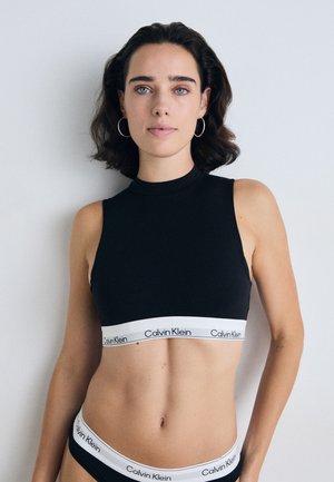 Kvinne med kort, mørkt hår iført svart Calvin Klein crop top og matchende undertøy, stående mot en ensfarget lys bakgrunn.