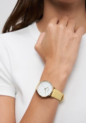Round wristwatch with a white face, gold hardware, and a soft yellow leather strap, worn on a lightly tanned arm against a white background.