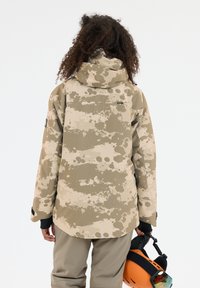 Beige camo waterdichte skijack met een capuchon, met een gladde textuur en subtiele aquarelachtige patronen; helm in de hand vastgehouden.